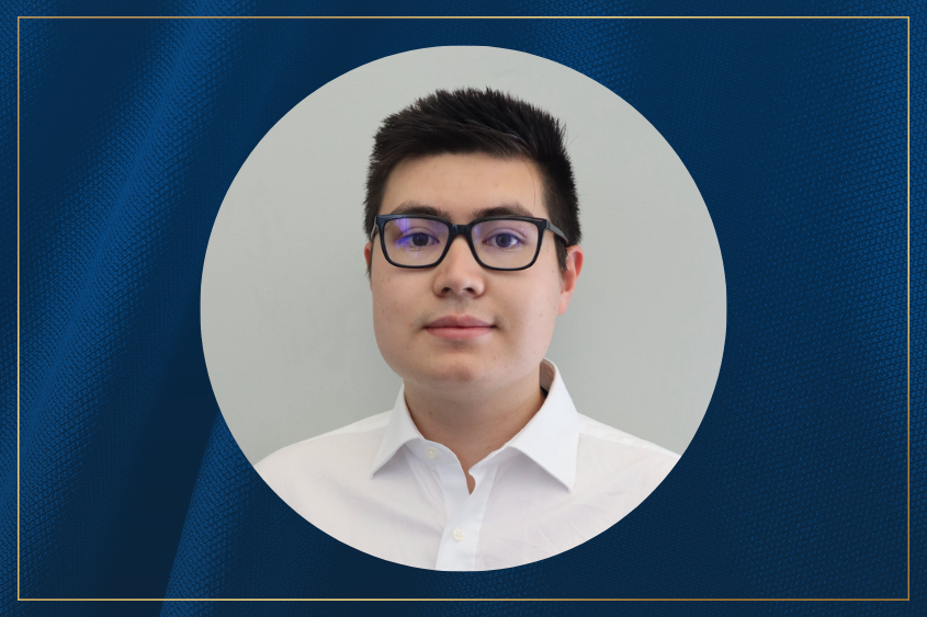 Portrait of a person with short dark hair, wearing glasses and a white collared shirt, standing against a plain light gray background.