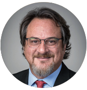 A man in a formal black suit and light blue shirt, wearing a red tie with a subtle pattern, poses against a neutral backdrop.